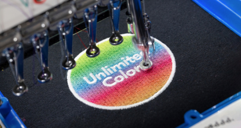 Close-up of an embroidery machine stitching a circular “Unlimited Colors” design with a vibrant gradient on black fabric.